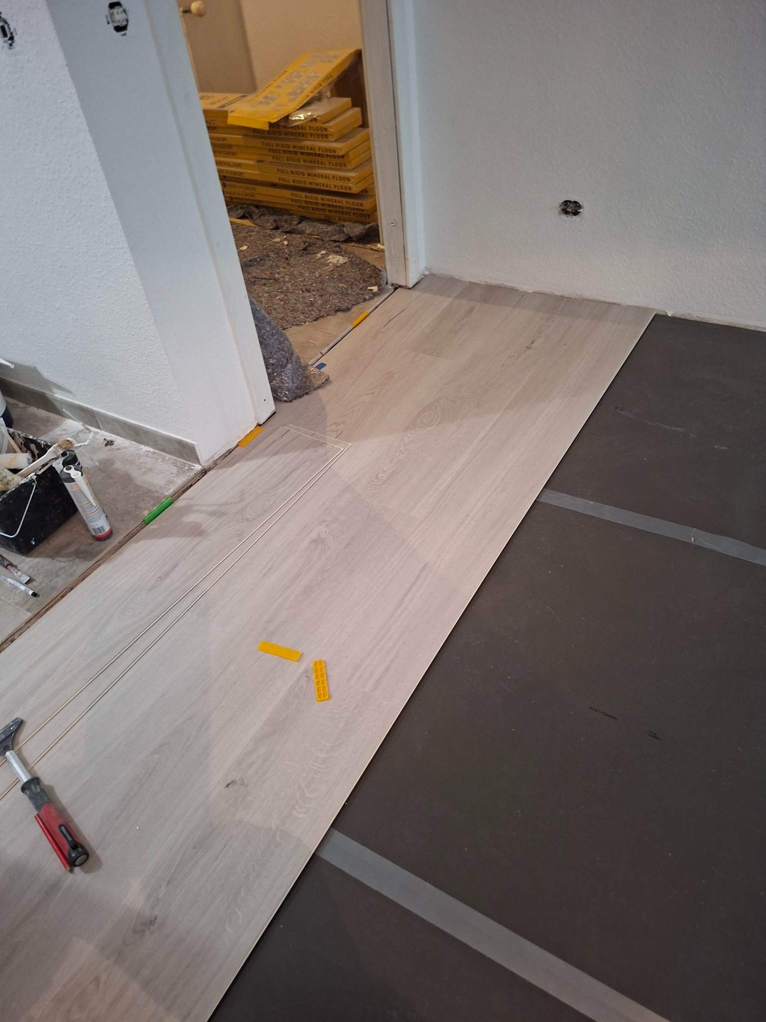 Układanie jasnych paneli podłogowych na czarnej macie izolacyjnej. Widoczne narzędzia i kliny dystansowe. W tle paczki paneli.