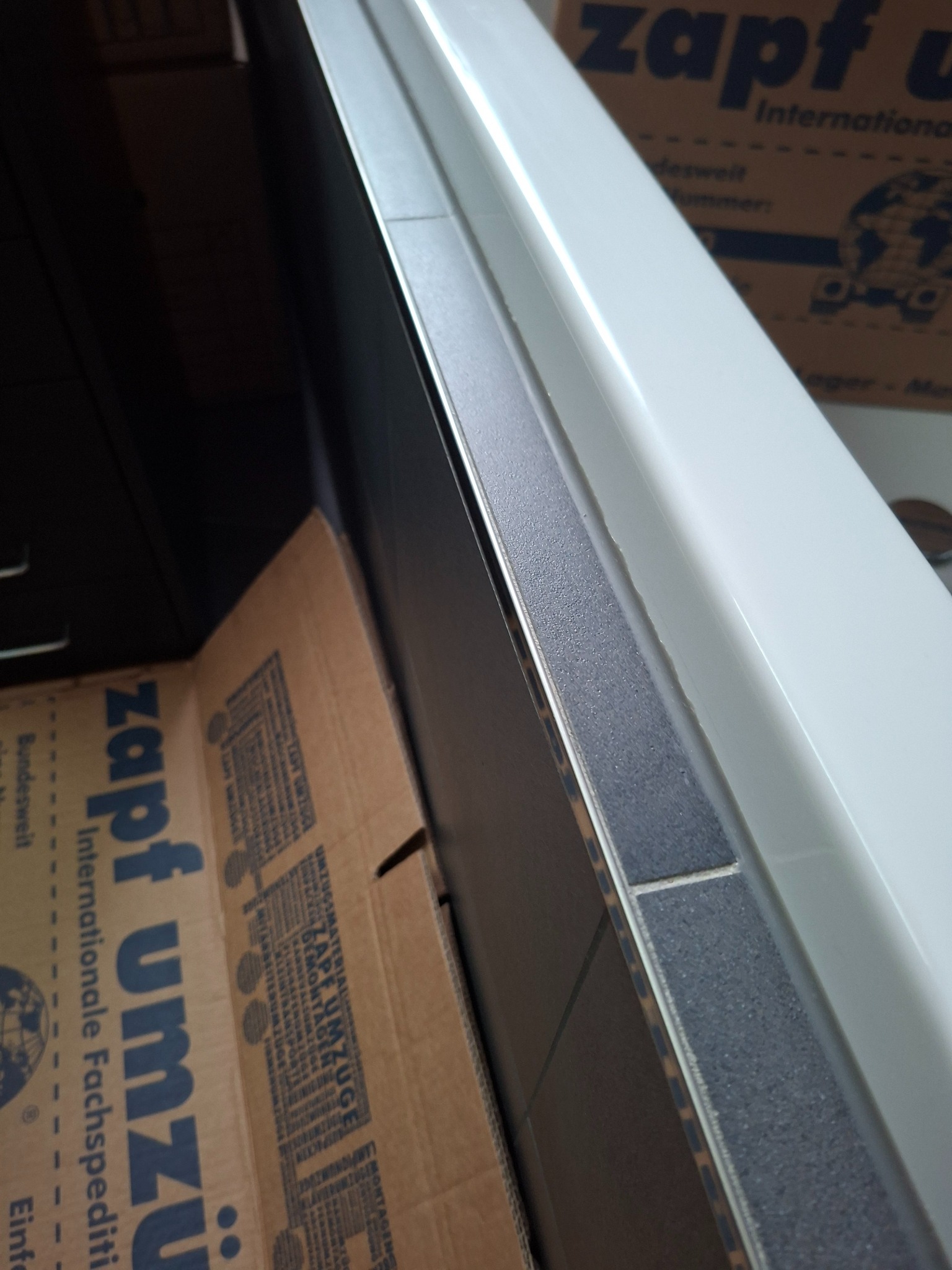 Szczegółowe ujęcie wykończenia krawędzi w łazience, z widocznymi szarymi płytkami i białą wanną, na tle kartonowych pudeł. Nietypowy kąt i bliskie ujęcie.