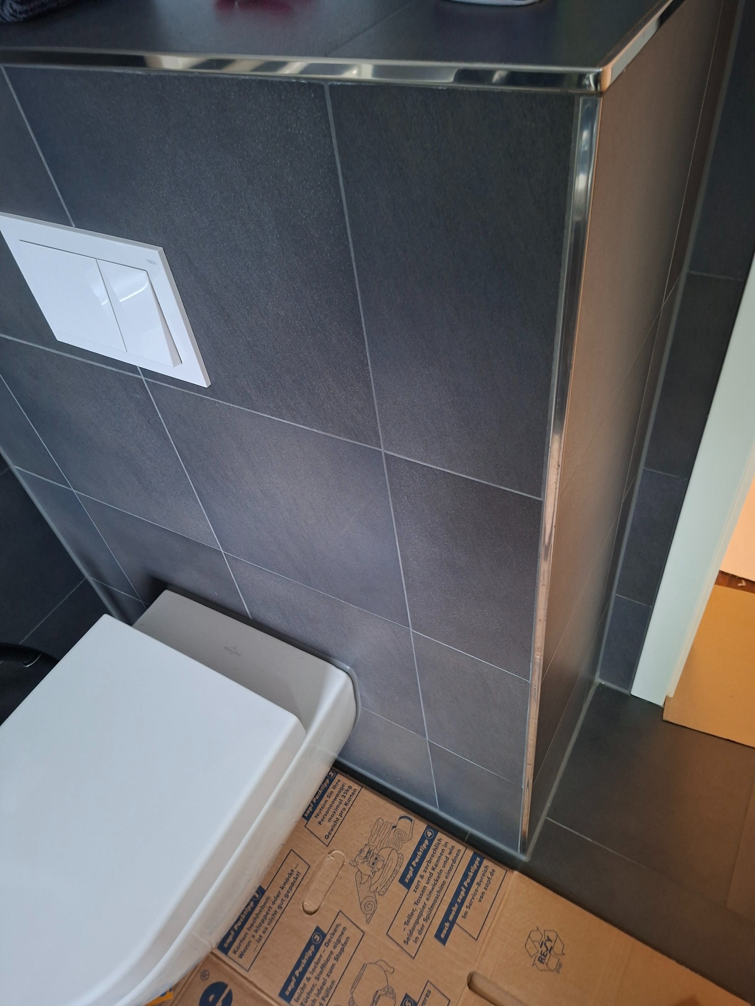 Nowoczesna toaleta w łazience wykończonej ciemnymi płytkami z metalowymi listwami. Widoczny biały przycisk spłuczki i fragment podłogi z kartonem.