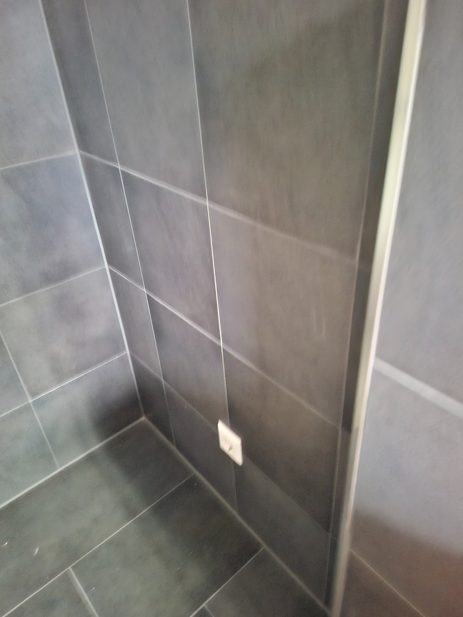 Szare, matowe płytki w kabinie prysznicowej z widocznym gniazdkiem elektrycznym i fragmentem ramy kabiny. Minimalistyczny design, nowoczesne wykończenie.