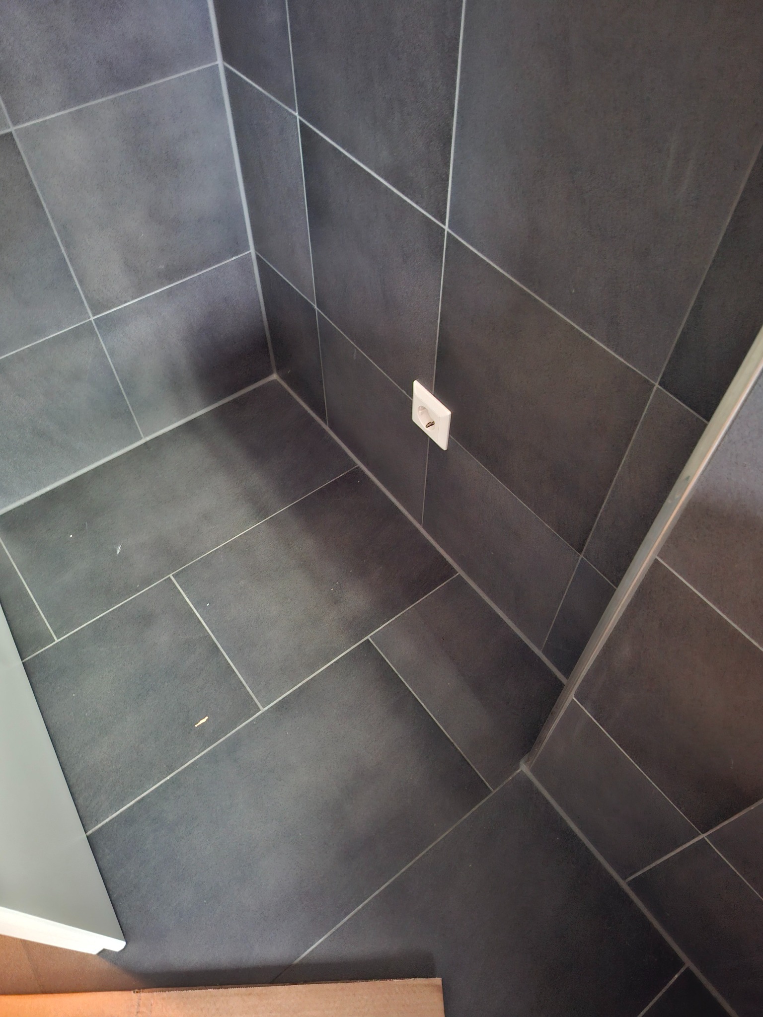 Ciemnoszare płytki podłogowe i ścienne ułożone w narożniku pomieszczenia, widoczne gniazdko elektryczne i fragment białej listwy przypodłogowej.