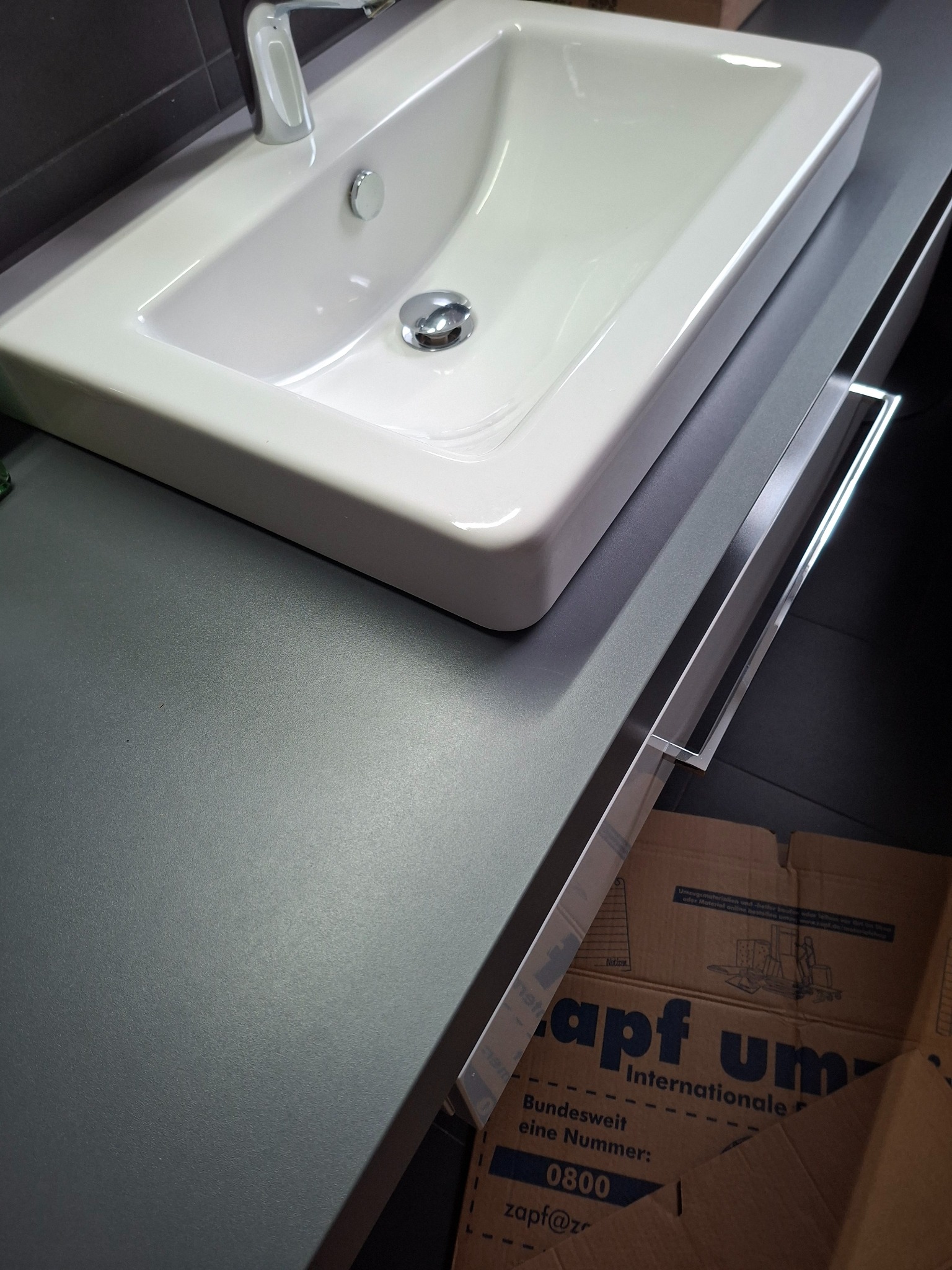 Biała umywalka z chromowaną baterią na szarym blacie szafki łazienkowej. Widoczny fragment kartonu z nadrukiem. Minimalistyczny design.