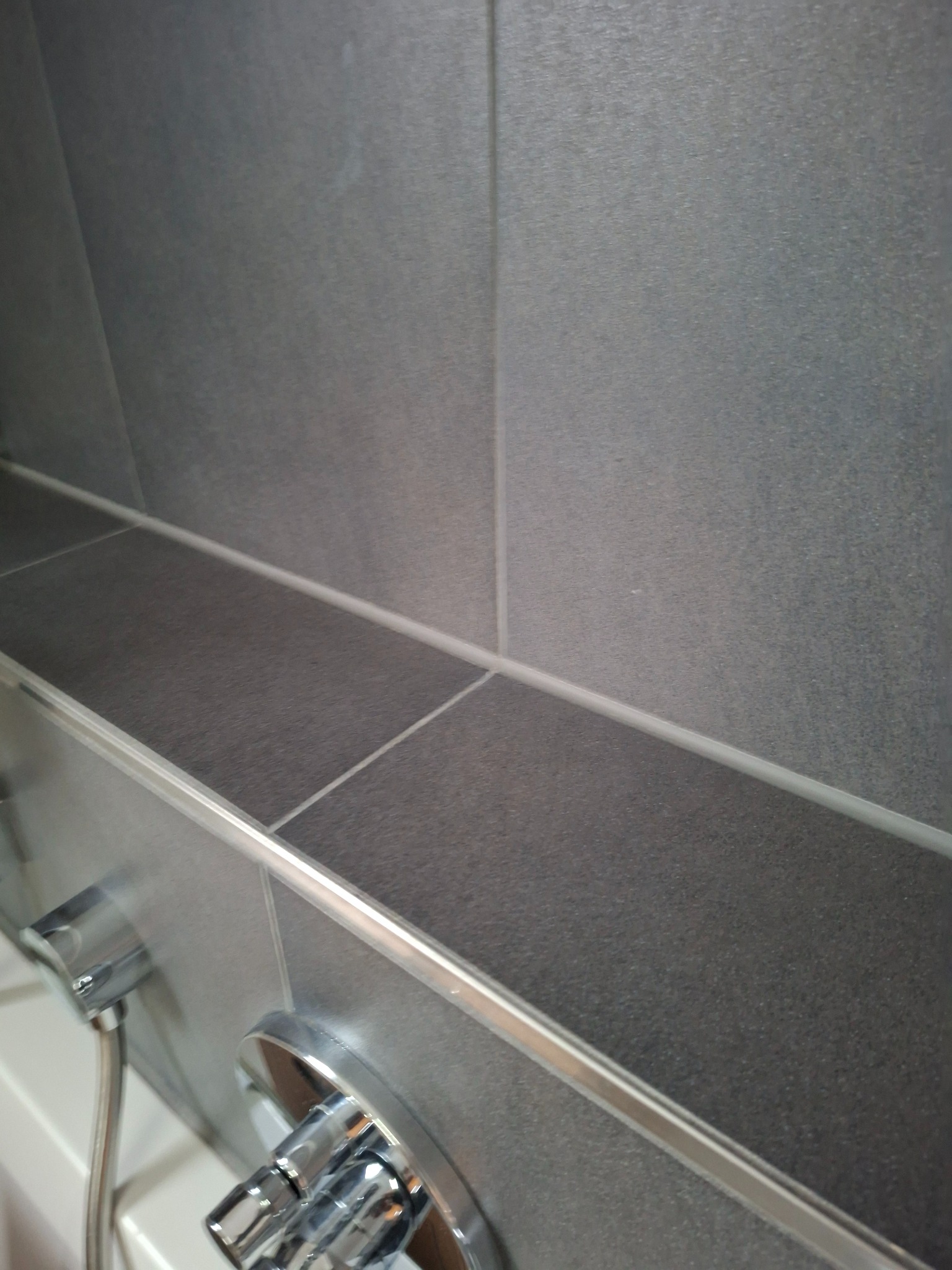 Szare, matowe płytki w łazience z półką i chromowaną baterią. Widoczne fugi i listwy wykończeniowe. Ujęcie z bliska, podkreślające detale.