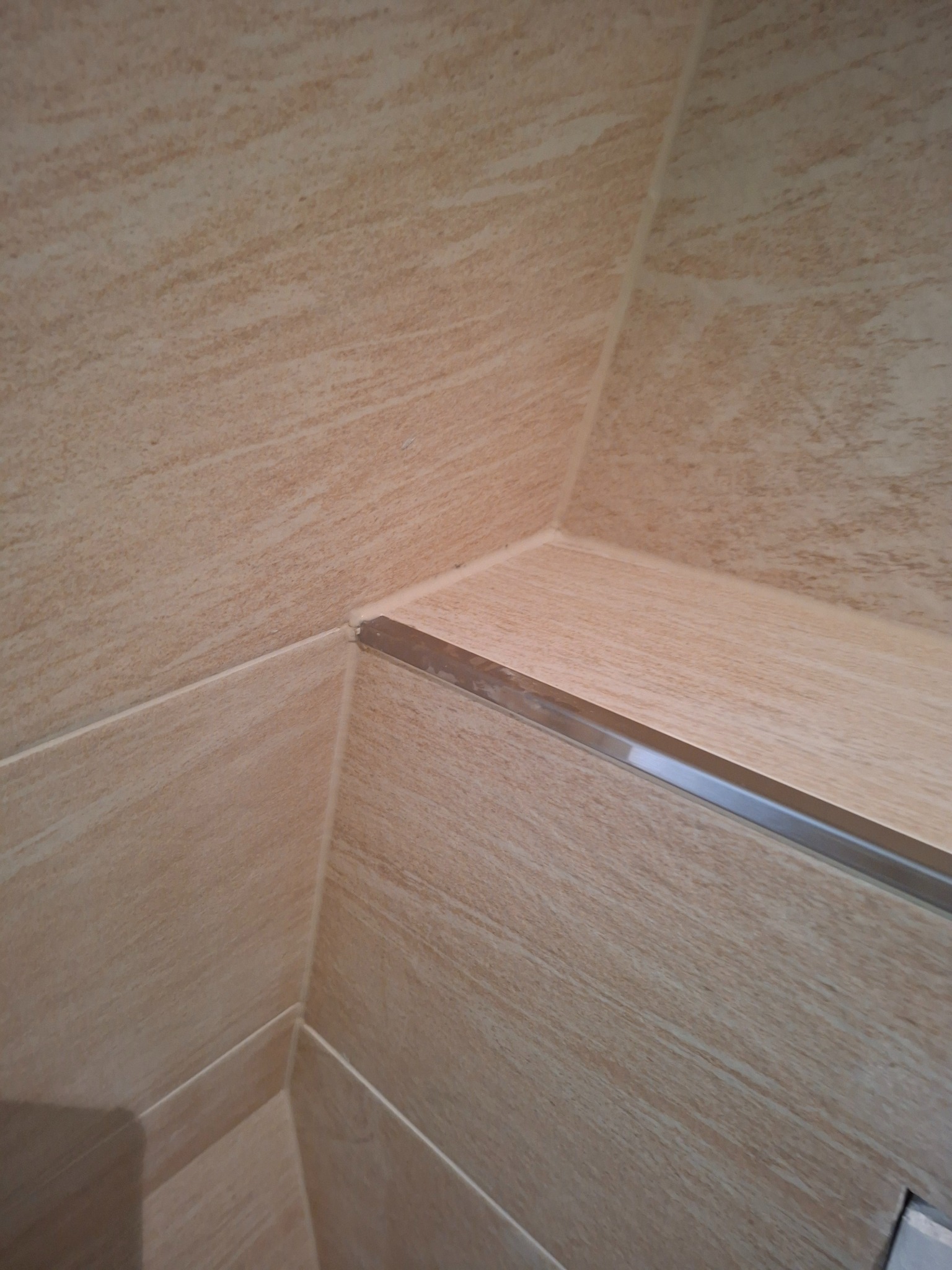 Beżowe płytki imitujące drewno, ułożone w rogu pomieszczenia, wykończone metalową listwą. Widoczne fugi i struktura płytek.