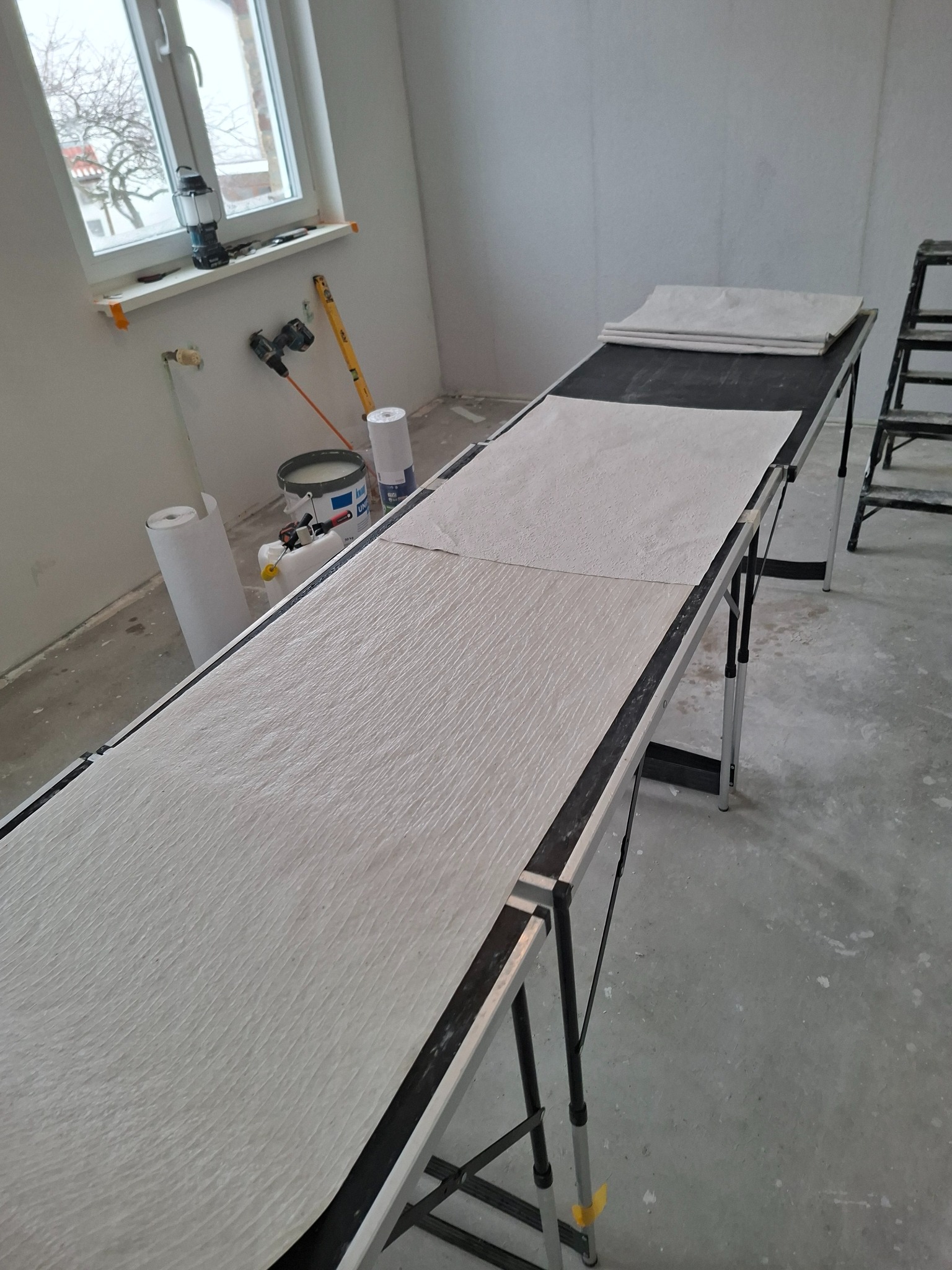 Przygotowanie tapety z włókna szklanego na długim stole roboczym w pomieszczeniu w trakcie remontu. Widoczne narzędzia i materiały malarskie.