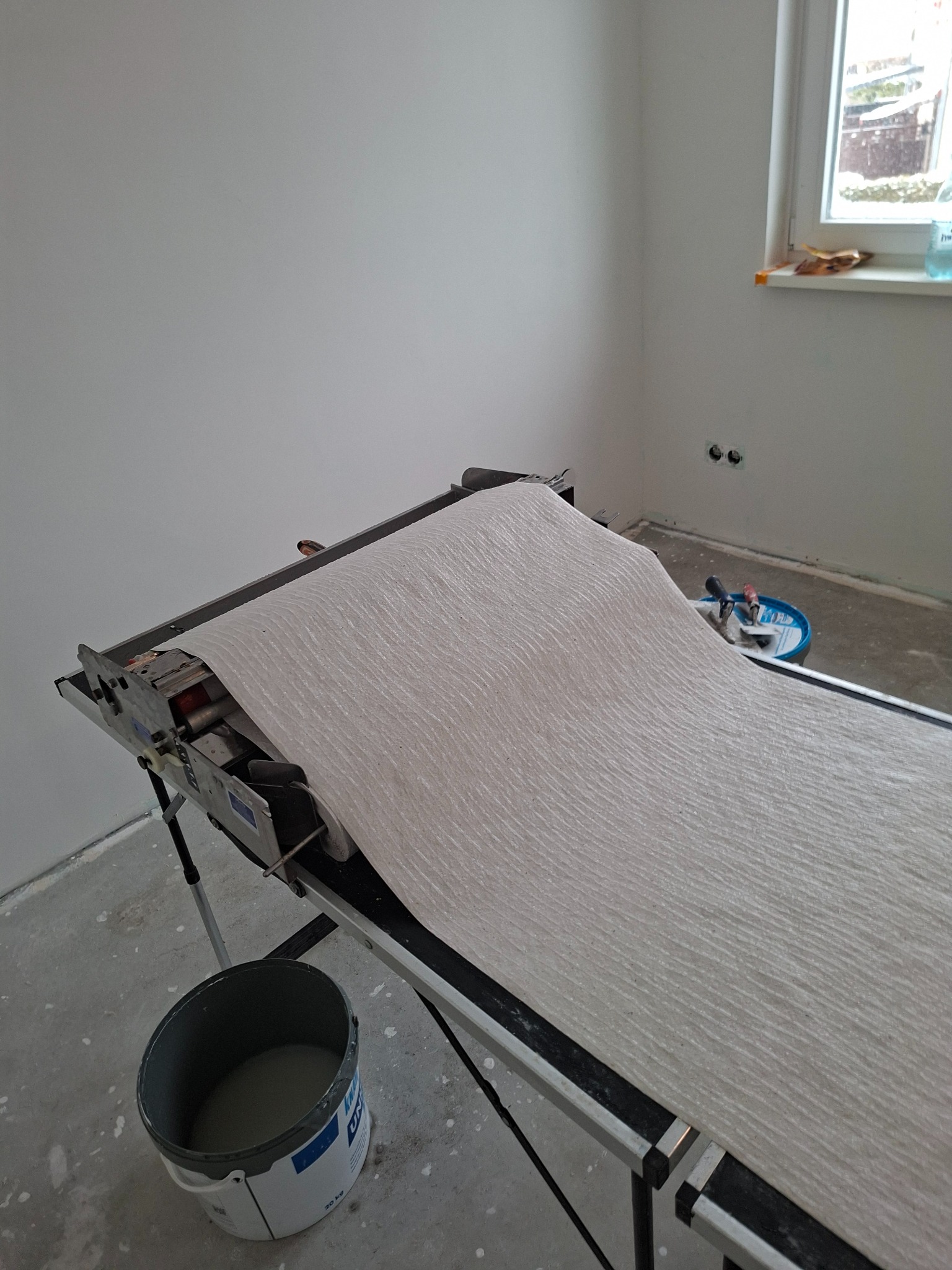 Tapeta na stole do tapetowania w jasnym wnętrzu. Widoczny kubeł z klejem i narzędzia. Przygotowanie do tapetowania ścian w pokoju z oknem.