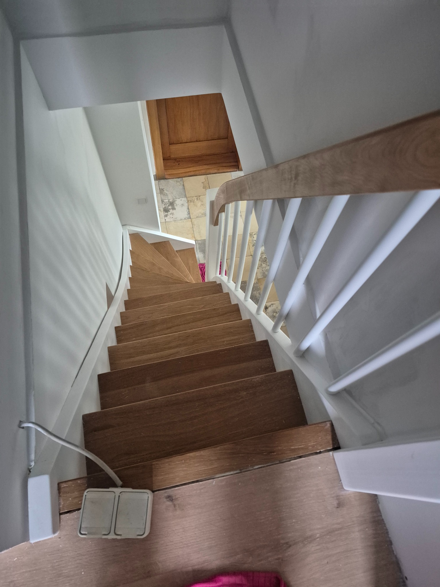 Widok z góry na drewniane schody z białymi balustradami w jasnym wnętrzu. U dołu widoczny fragment podłogi i włącznik światła.