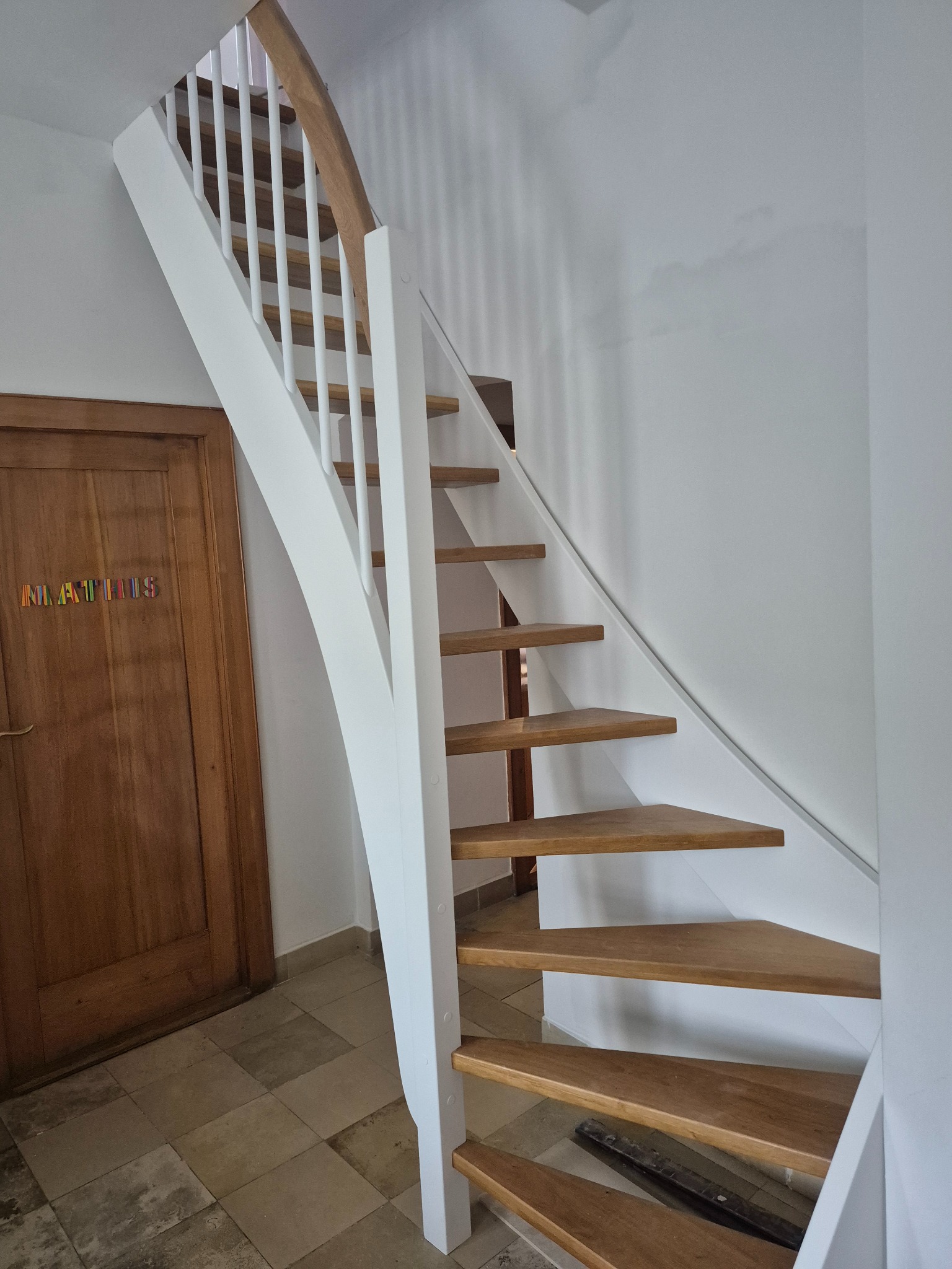 Drewniane schody z białą balustradą w jasnym wnętrzu. Widoczna drewniana podłoga i drzwi z kolorowym napisem. Ujęcie z dołu, podkreślające konstrukcję schodów.