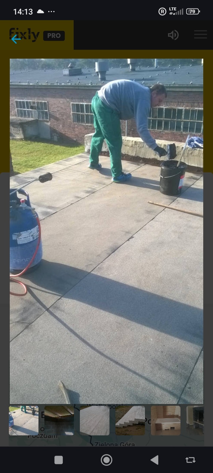 Pracownik w trakcie hydroizolacji dachu płaskiego. Widoczny zbiornik z gazem, wiadro z masą bitumiczną i wałek. W tle budynek przemysłowy z oknami.