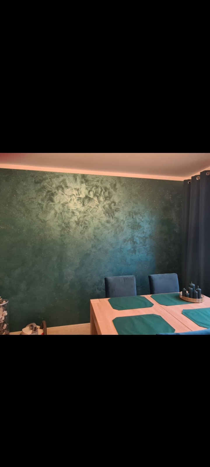 Dekoracyjna ściana w odcieniach zieleni z efektem marmuru, oświetlona od góry. W tle stół z zielonymi podkładkami i krzesła w podobnym odcieniu.