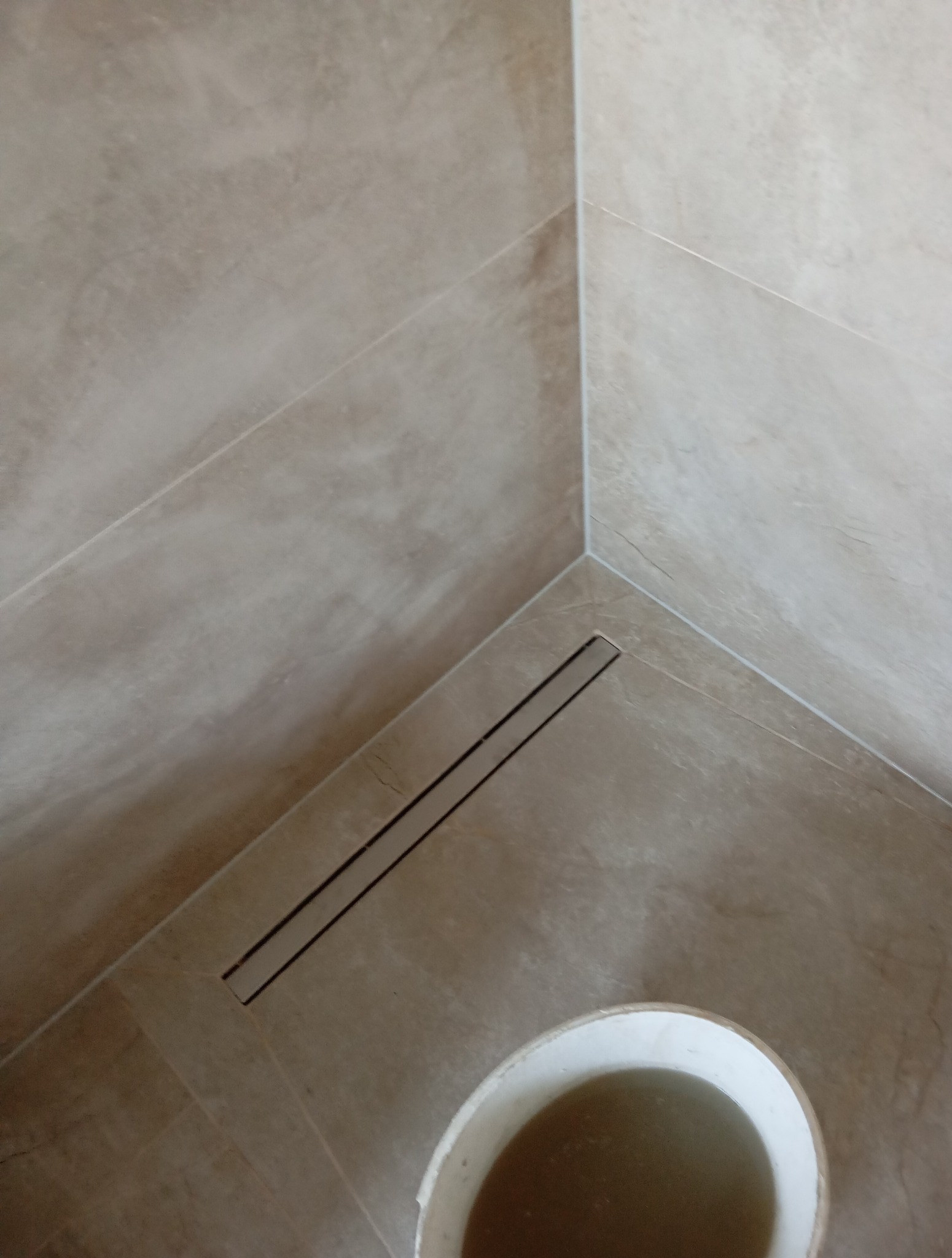 Beżowe płytki w kabinie prysznicowej z odpływem liniowym. Widoczny fragment wiadra z wodą. Ujęcie z góry, podkreślające detale wykończenia i estetykę.