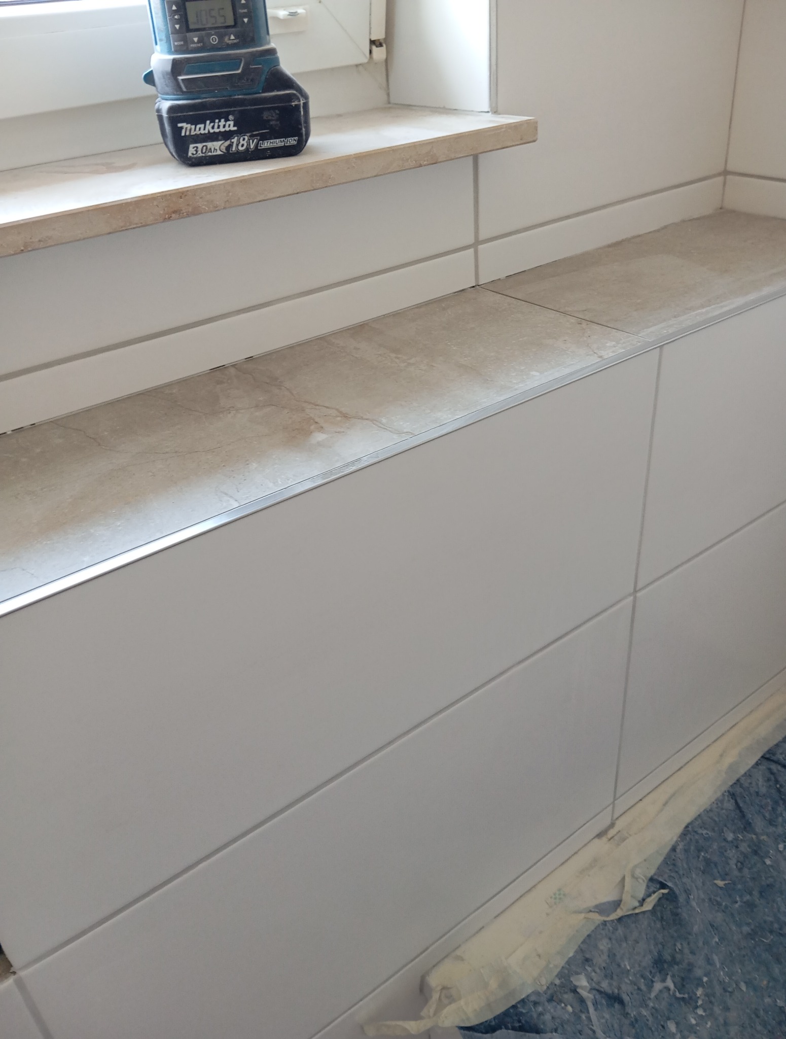 Wykończona wnęka okienna z płytkami w odcieniach bieli i beżu, z widocznym narzędziem akumulatorowym Makita na parapecie. Detal wykończenia wnętrza po remoncie.