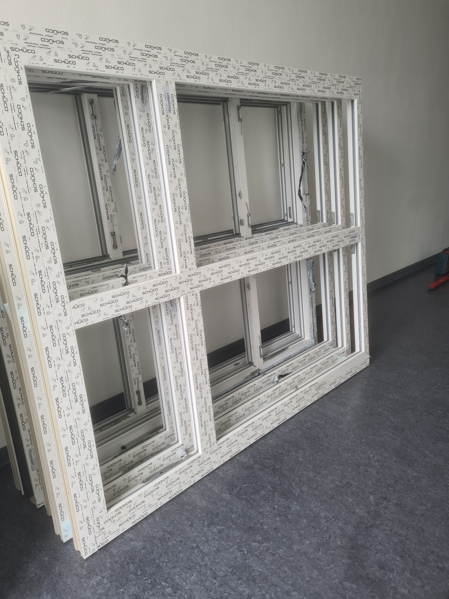 Białe ramy okienne Schüco, ułożone jedna na drugiej, czekają na montaż. Widoczne detale profili i mechanizmy okienne. Szary podkład i jasne ściany w tle.