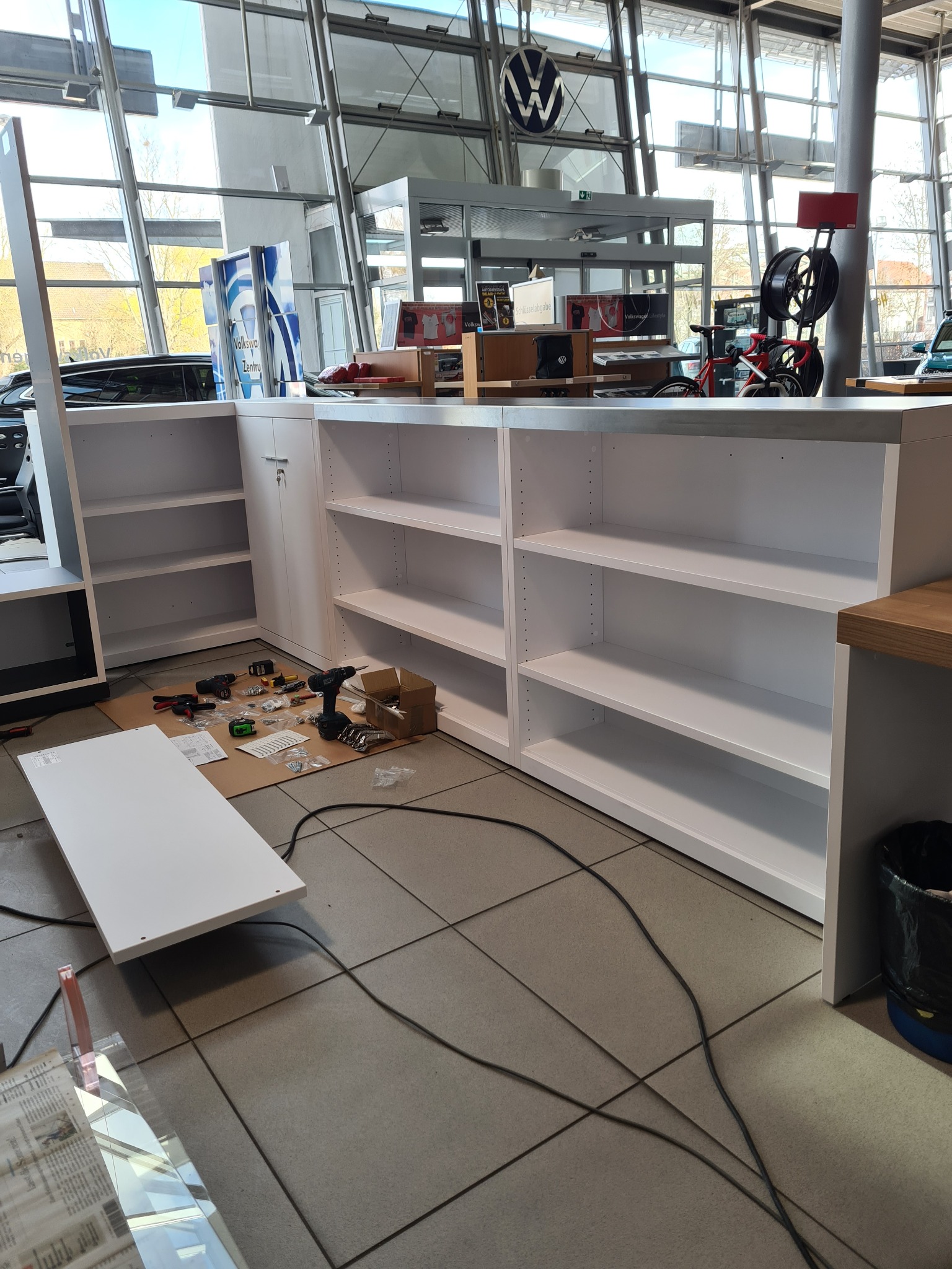 Białe regały z półkami w trakcie montażu w salonie samochodowym. Widoczne narzędzia i elementy montażowe. Jasne wnętrze z dużymi oknami i logo marki.