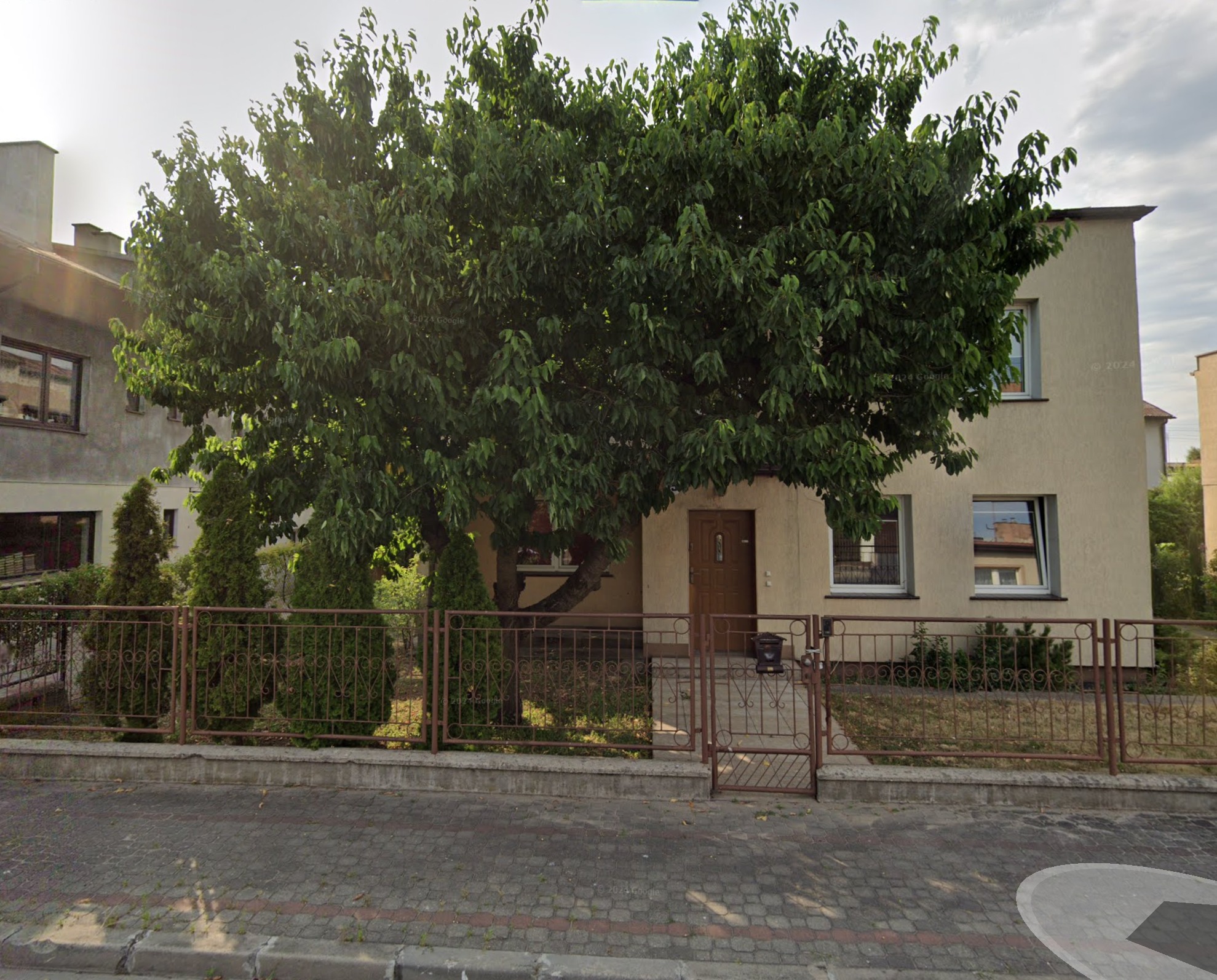 Dwupiętrowy budynek z beżową elewacją, częściowo zasłonięty przez rozłożyste drzewo liściaste i ozdobne tuje, ogrodzony metalowym płotem z furtką.