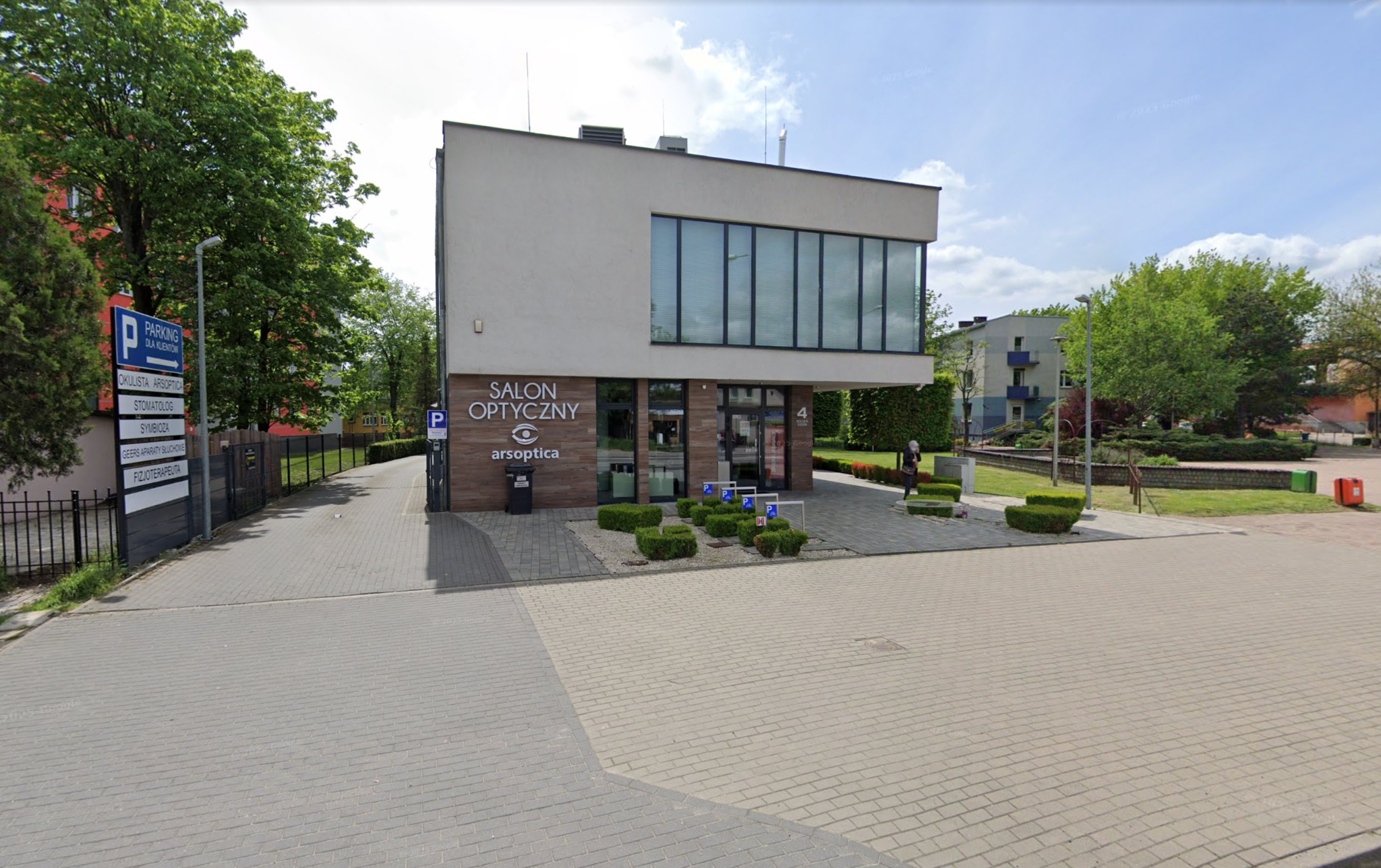 Nowoczesny budynek salonu optycznego z dużymi oknami i logo 'Salon Optyczny arsoptica'. Przed budynkiem zadbany chodnik i zieleń. Tablica informacyjna o parkingu dla klientów.
