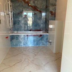 Babylon Mateusz Pytka - Wykończona łazienka z marmurowymi płytkami w odcieniach błękitu i beżu. Widoczna wanna obudowana płytkami, grzejnik drabinkowy i fragment toalety. W tle okno z widokiem.