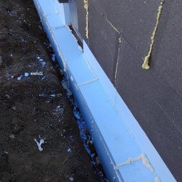 Babylon Mateusz Pytka - Ocieplenie fundamentów budynku: białe płyty styropianowe przytwierdzone do ściany z czarnego styropianu, wypełnione pianką montażową, widok z bliska na wykop.