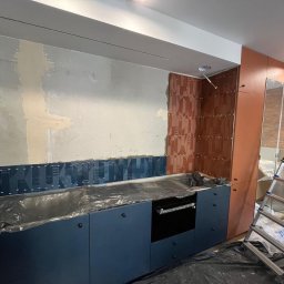 Babylon Mateusz Pytka - Montaż dekoracyjnych płytek ściennych w kuchni. Widoczne fragmenty ściany przed i po położeniu płytek. Zabezpieczone folią meble kuchenne i drabina w tle.