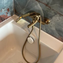 Babylon Mateusz Pytka - Bateria wannowa w kolorze starego złota, zamontowana na białej wannie z odpływem. W tle marmurowe płytki w odcieniach błękitu i brązu. Detal wykończenia łazienki.