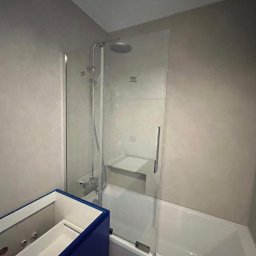 Babylon Mateusz Pytka - Nowoczesna kabina prysznicowa ze szklaną ścianką nad wanną, widoczna armatura i fragment niebieskiej szafki pod umywalkę. Minimalistyczny design łazienki.