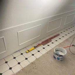 Babylon Mateusz Pytka - Ściana z dekoracyjnymi panelami i poziomymi listwami, poziomice na podłodze z czarno-białej mozaiki, wiadro z farbą i lampa budowlana. Detal wykończenia wnętrza.