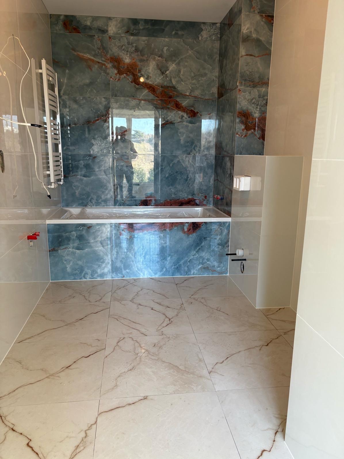 Wykończona łazienka z marmurowymi płytkami w odcieniach błękitu i beżu. Widoczna wanna obudowana płytkami, grzejnik drabinkowy i fragment toalety. W tle okno z widokiem.