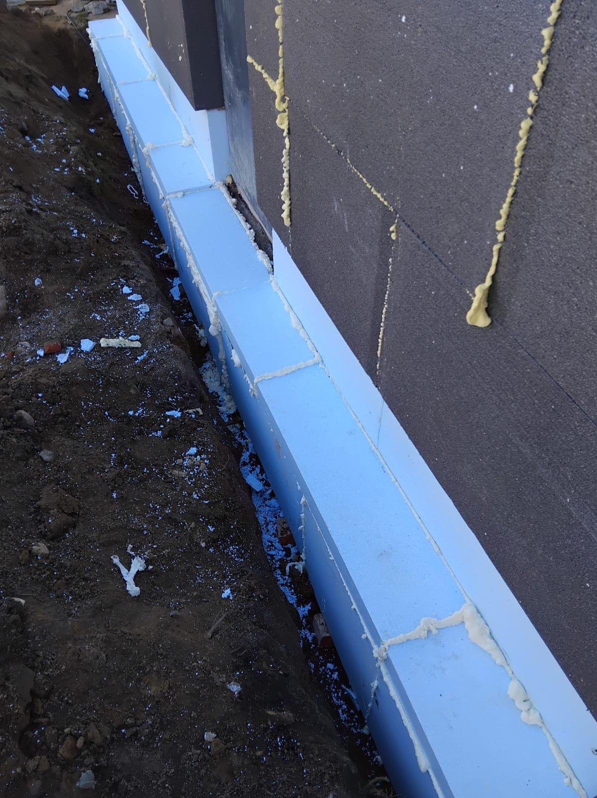 Ocieplenie fundamentów budynku: białe płyty styropianowe przytwierdzone do ściany z czarnego styropianu, wypełnione pianką montażową, widok z bliska na wykop.