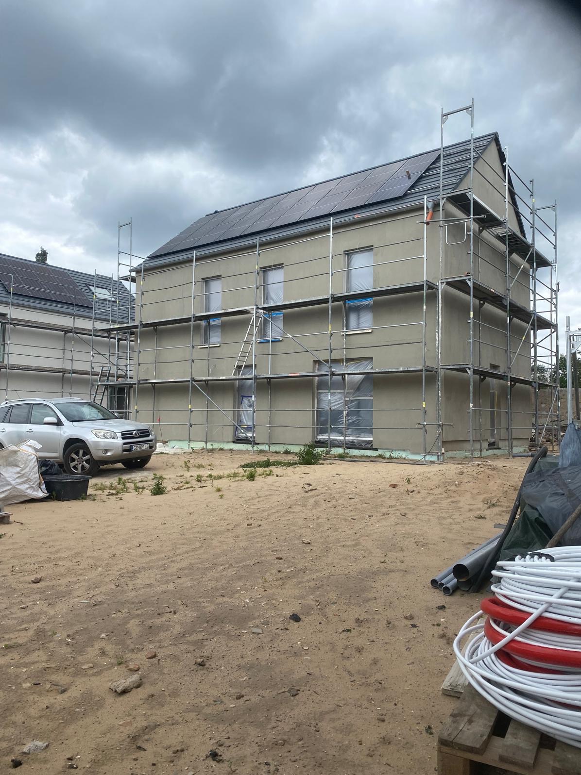 Dom w trakcie szpachlowania elewacji, otoczony rusztowaniami. Okna zabezpieczone folią. Na dachu panele słoneczne. Obok zaparkowany srebrny samochód.