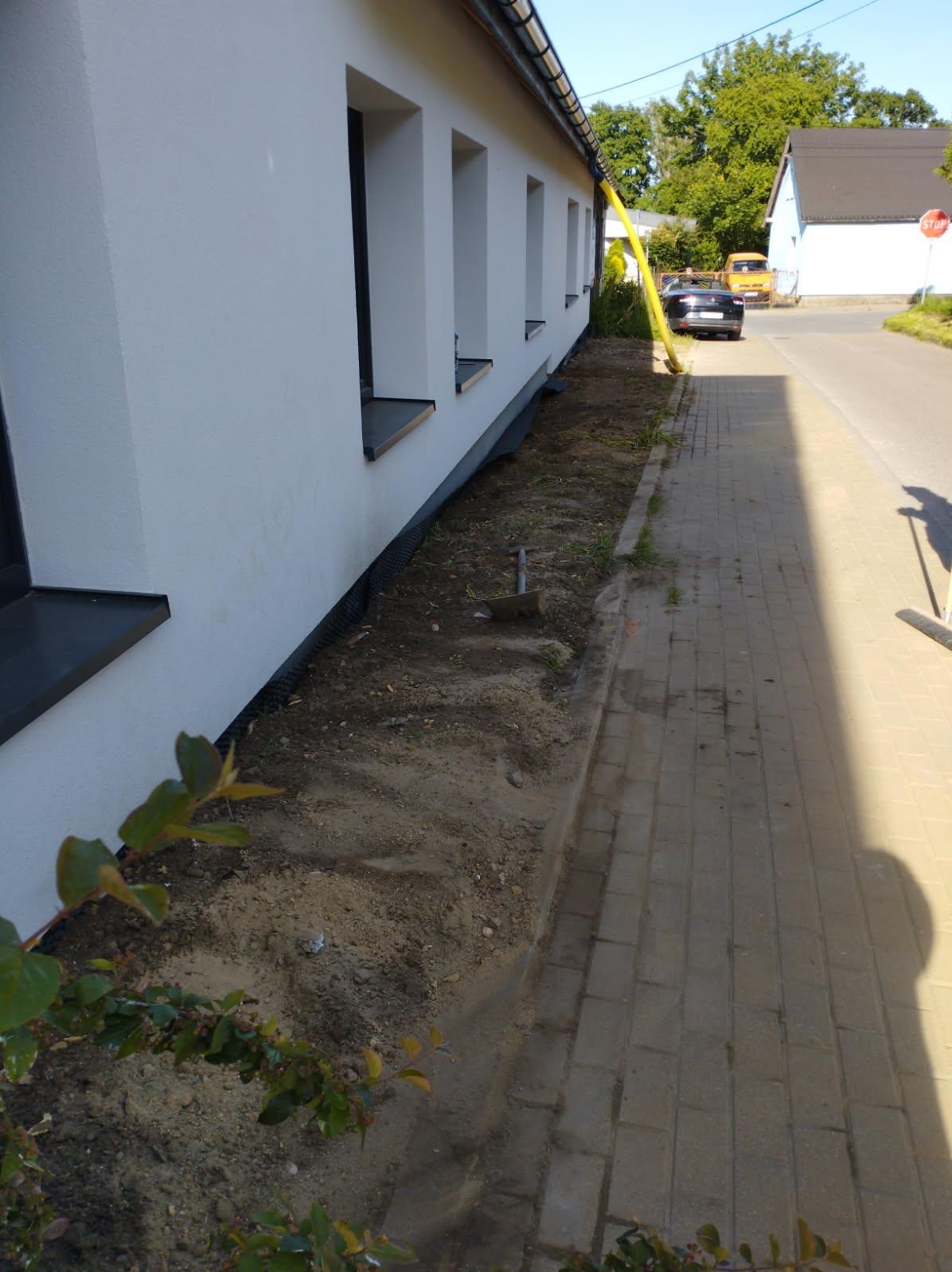 Ocieplony budynek z nową elewacją, widoczna warstwa izolacyjna przy gruncie i przygotowany teren pod nasadzenia. Świeżo pomalowana fasada w jasnym odcieniu.