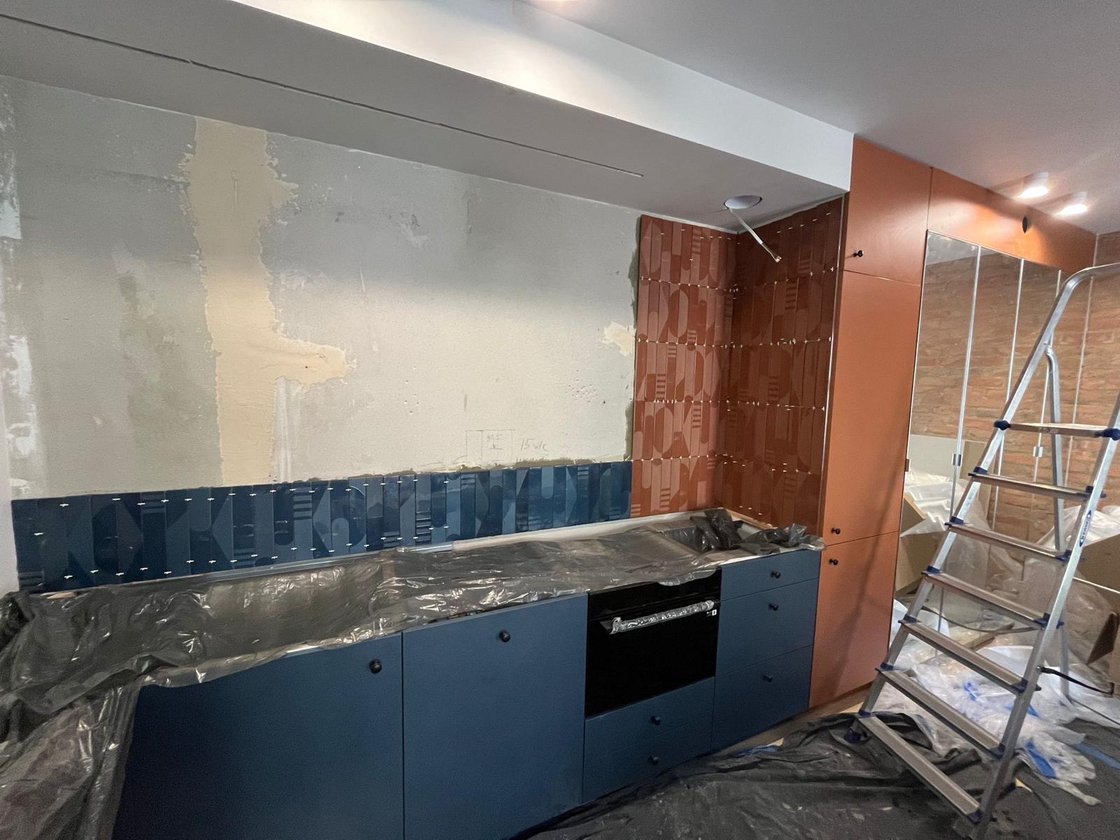 Montaż dekoracyjnych płytek ściennych w kuchni. Widoczne fragmenty ściany przed i po położeniu płytek. Zabezpieczone folią meble kuchenne i drabina w tle.