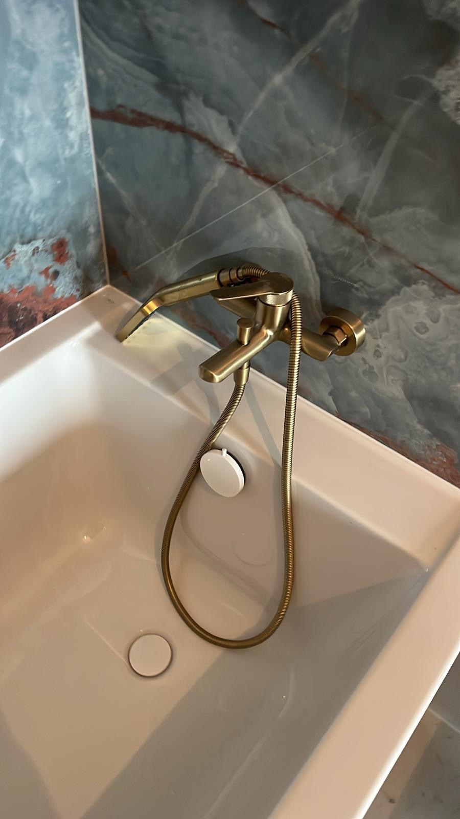 Bateria wannowa w kolorze starego złota, zamontowana na białej wannie z odpływem. W tle marmurowe płytki w odcieniach błękitu i brązu. Detal wykończenia łazienki.