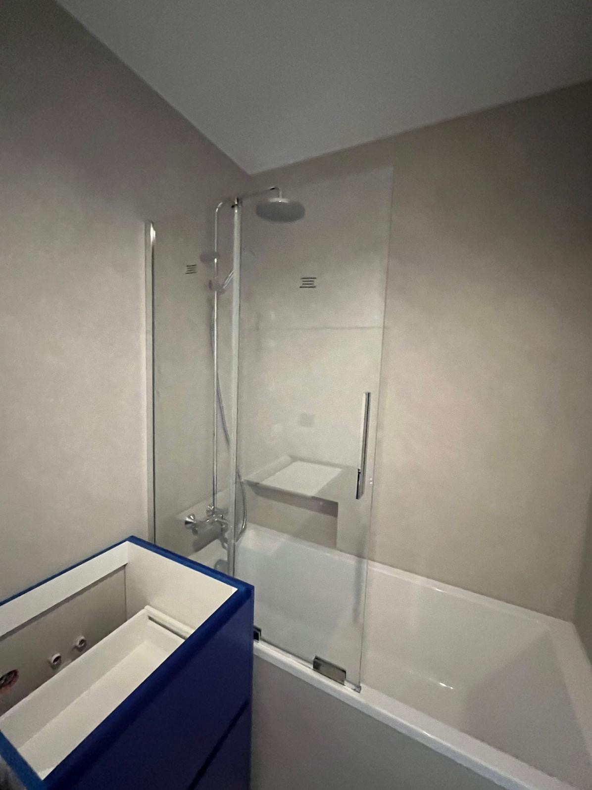 Nowoczesna kabina prysznicowa ze szklaną ścianką nad wanną, widoczna armatura i fragment niebieskiej szafki pod umywalkę. Minimalistyczny design łazienki.