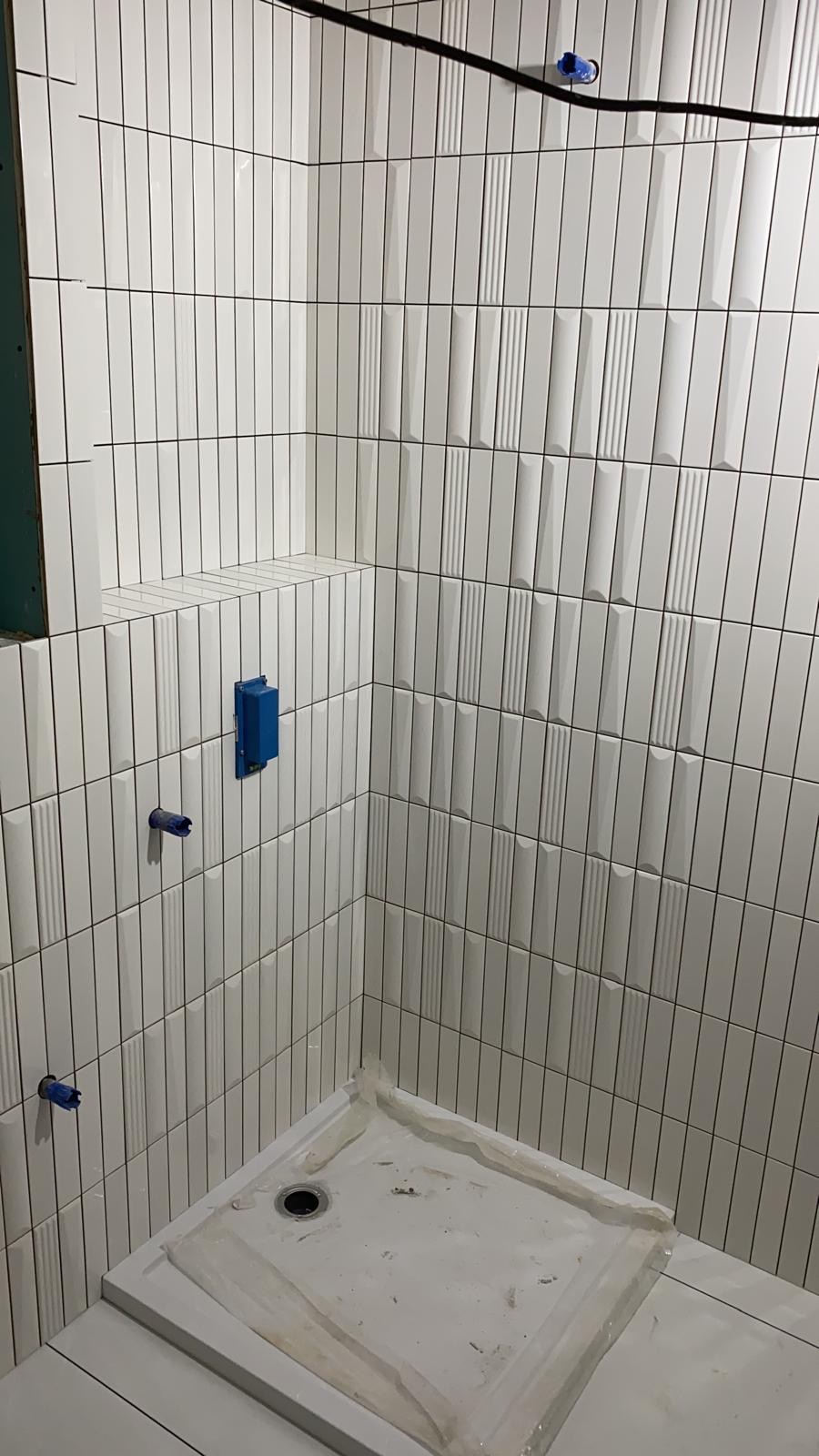 Białe, pionowo ułożone płytki w łazience, z widocznym brodzikiem i niebieskimi elementami instalacji. Częściowo wykończona ściana z płyt karton-gips.