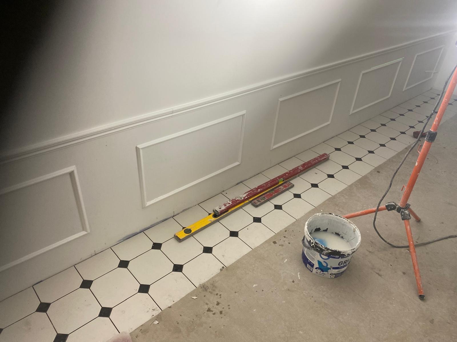 Ściana z dekoracyjnymi panelami i poziomymi listwami, poziomice na podłodze z czarno-białej mozaiki, wiadro z farbą i lampa budowlana. Detal wykończenia wnętrza.