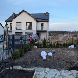 MiH Service - Nowoczesny dom z panelami słonecznymi widziany z ogrodu z młodymi tujami i dekoracyjnymi kulami. W tle las i błękitne niebo o zmierzchu.