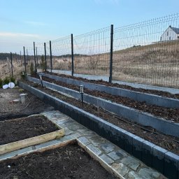 MiH Service - Przygotowane grządki warzywne z obrzeżami i ścieżką z kamienia, oświetlone lampkami solarnymi, ogrodzone siatką. Widok na wzgórze i dom w tle.