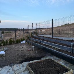 MiH Service - Ogród z podwyższonymi grządkami z szarej kostki, oświetleniem solarnym i drewnianą skrzynią na ziemię, otoczony siatką ogrodzeniową na tle pola i lasu.