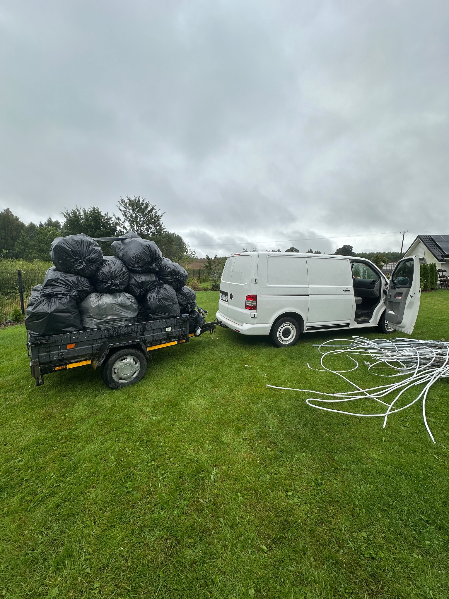 Przyczepka wypełniona workami na śmieci, zaparkowana obok białego busa z otwartymi drzwiami na trawniku. W tle dom z panelami słonecznymi i pochmurne niebo.