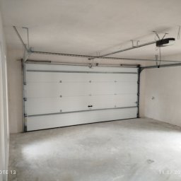 Mariusz Sandacz - Biała segmentowa brama garażowa z automatycznym napędem, widoczna od wewnątrz, zamontowana w surowym garażu z betonową posadzką i otynkowanymi ścianami.