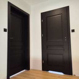 Mariusz Sandacz - Dwa czarne, eleganckie skrzydła drzwiowe z frezowanymi panelami, zamontowane w białym wnętrzu. Widoczna drewniana podłoga i czarne włączniki światła.