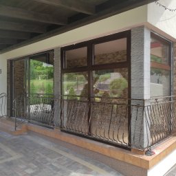 Mariusz Sandacz - Zadaszony taras z brązowymi oknami i ozdobną, metalową balustradą. Widoczne płytki imitujące kamień na elewacji i kostka brukowa na podłodze tarasu.