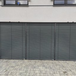 Mariusz Sandacz - Zewnętrzne, grafitowe żaluzje fasadowe zamontowane na budynku z dwoma oknami. Widoczny fragment drewnianej elewacji i kostka brukowa.