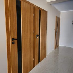 Mariusz Sandacz - Nowoczesne drzwi wewnętrzne z okleiną drewnopodobną i czarnymi wstawkami, widoczne trzy sztuki w korytarzu z jasną podłogą i ścianami.