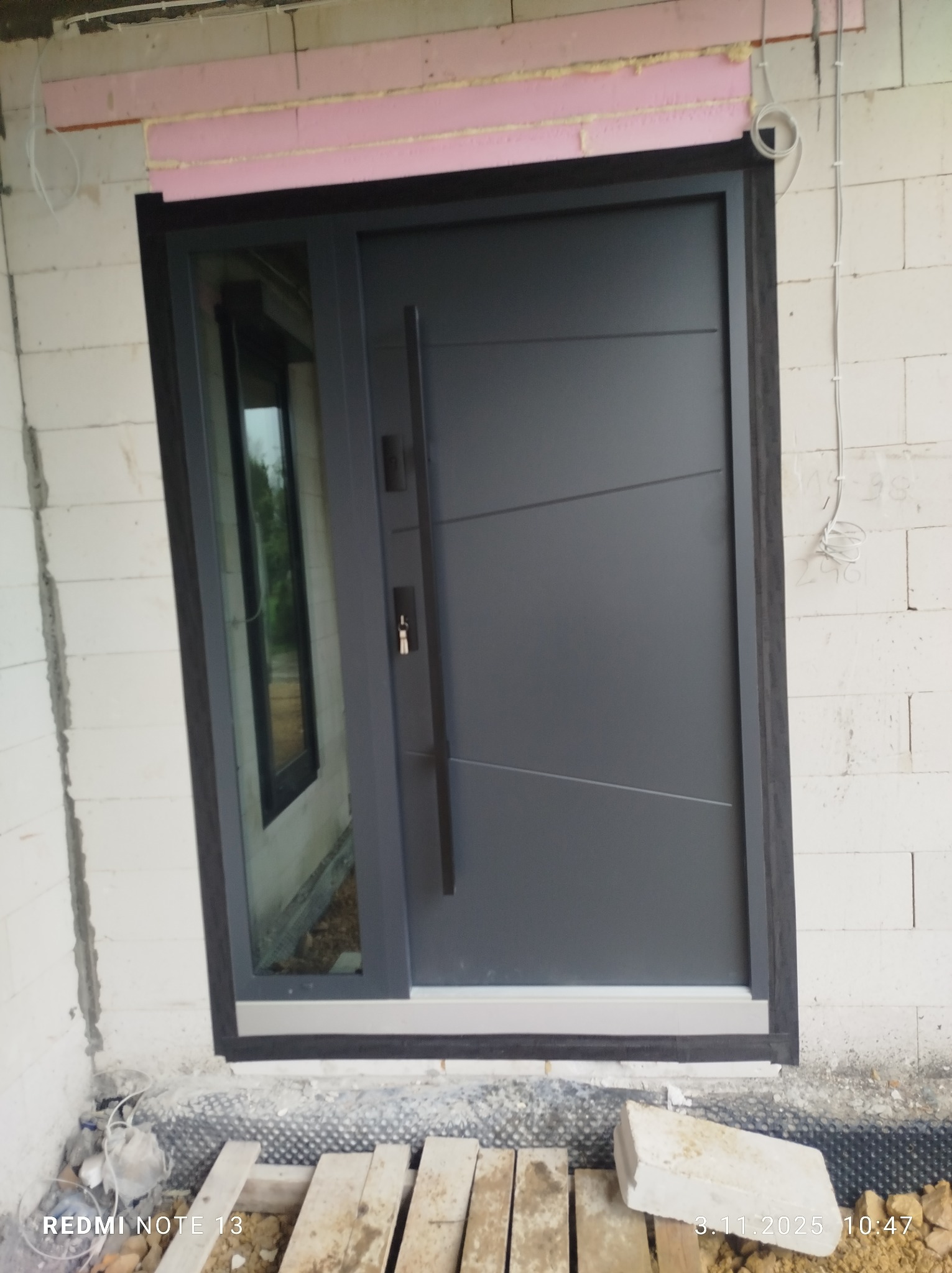 Nowoczesne, grafitowe drzwi zewnętrzne z przeszkleniem bocznym, zamontowane w surowym murze z widocznym ociepleniem. Minimalistyczny design, długa klamka.