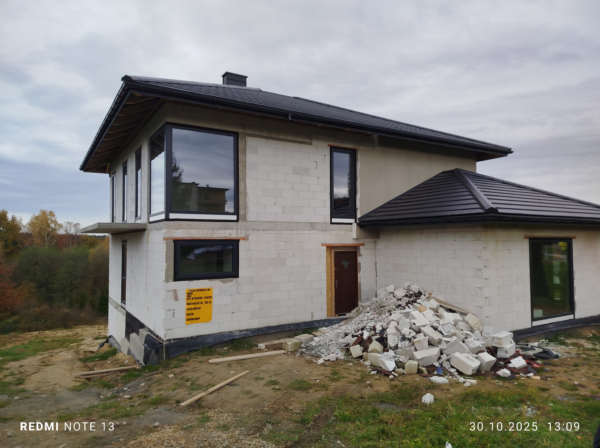 Surowy stan domu w budowie z czarnymi oknami i drzwiami, widoczne bloczki i gruz. Tablica informacyjna budowy. Pochmurne niebo w tle.