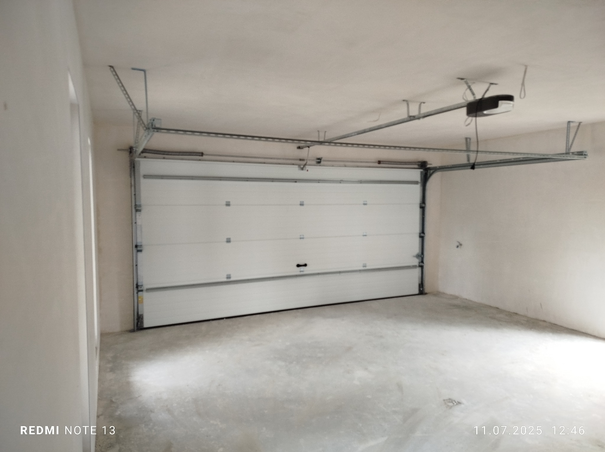 Biała segmentowa brama garażowa z automatycznym napędem, widoczna od wewnątrz, zamontowana w surowym garażu z betonową posadzką i otynkowanymi ścianami.