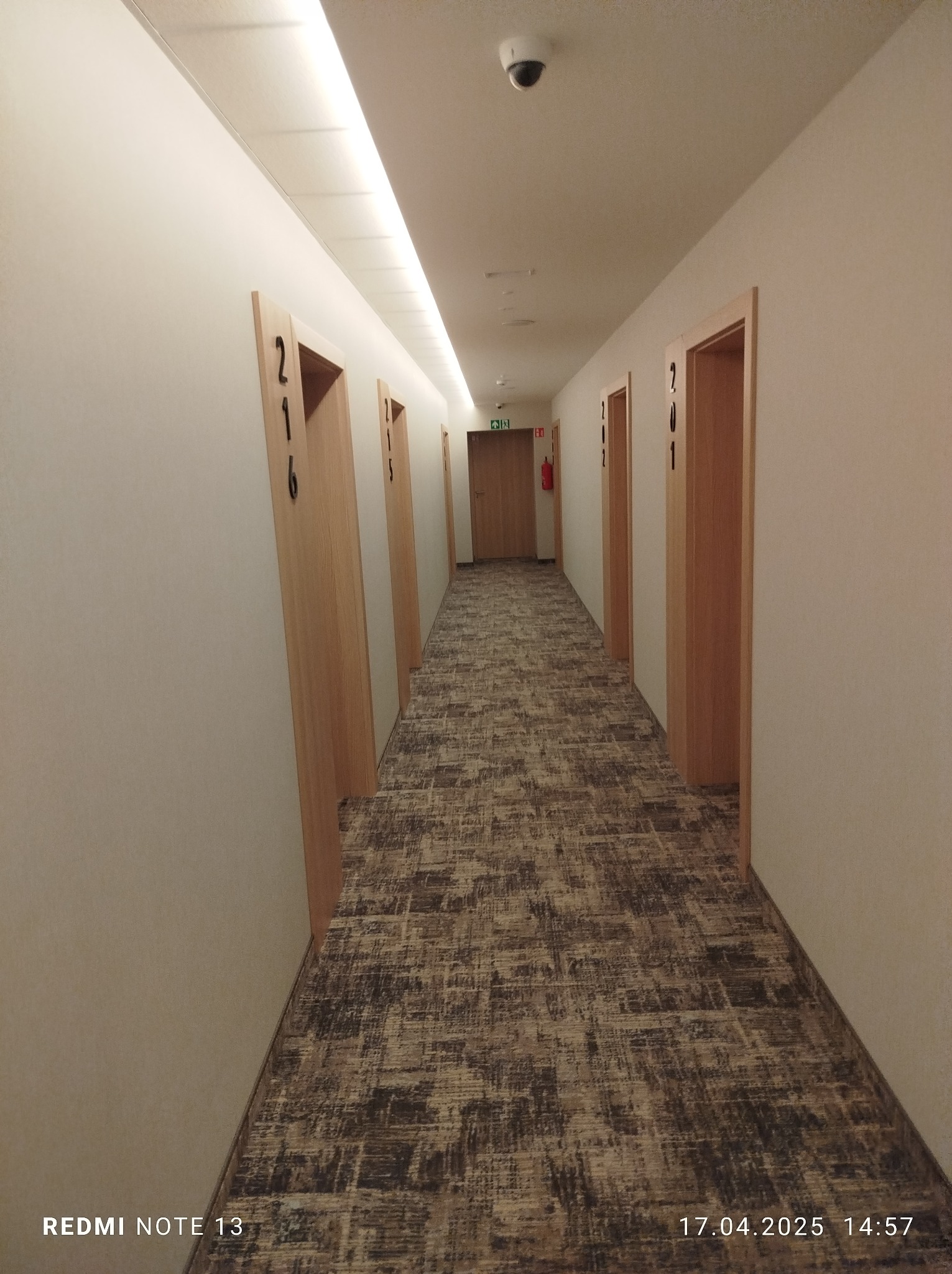 Długi korytarz hotelowy z drewnianymi drzwiami o numerach 216 i 201, wyłożony wzorzystą wykładziną. Widoczny system oświetlenia sufitowego i kamera monitoringu.