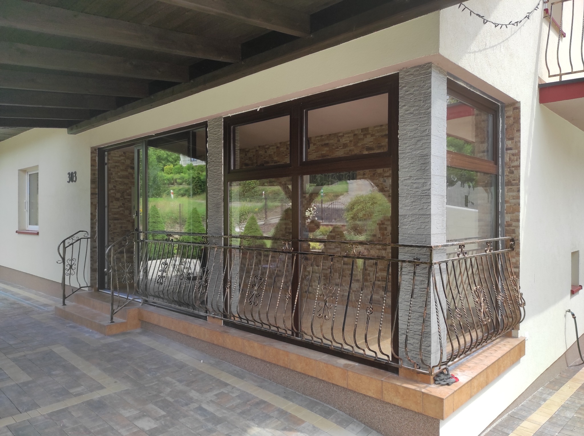 Zadaszony taras z brązowymi oknami i ozdobną, metalową balustradą. Widoczne płytki imitujące kamień na elewacji i kostka brukowa na podłodze tarasu.