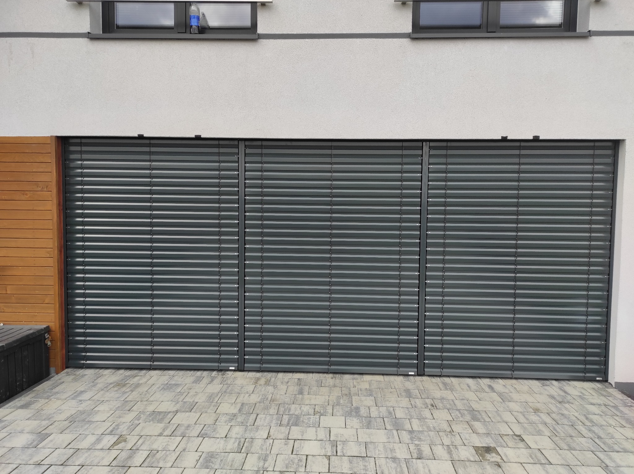 Zewnętrzne, grafitowe żaluzje fasadowe zamontowane na budynku z dwoma oknami. Widoczny fragment drewnianej elewacji i kostka brukowa.